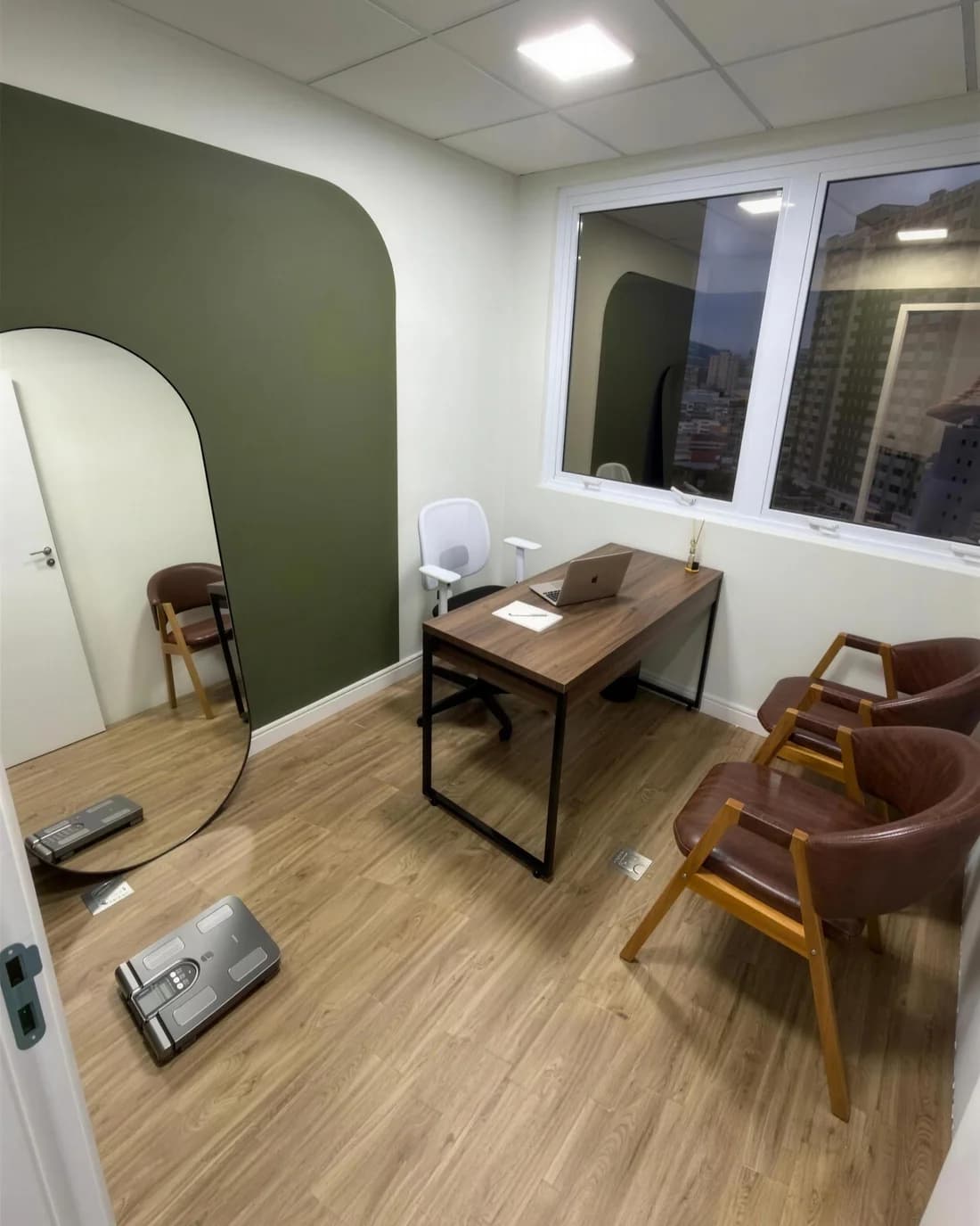 Sala de atendimento do consultório: parede oliva, mesa de madeira, poltronas e balança de bioimpedância no piso